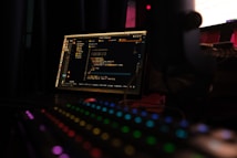 A dimly lit room features a laptop displaying code on its screen, implying a coding environment. In the foreground, a mechanical keyboard with colorful RGB lighting enhances the tech-focused setting.