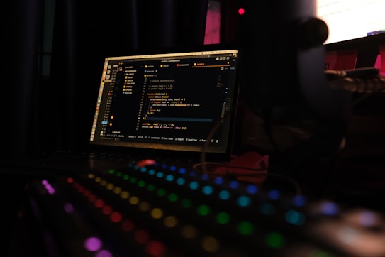 A dimly lit room features a laptop displaying code on its screen, implying a coding environment. In the foreground, a mechanical keyboard with colorful RGB lighting enhances the tech-focused setting.