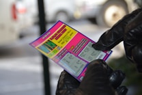 A person wearing black gloves is holding a colorful lottery scratch-off ticket. The ticket includes information about a $1,000,000 guaranteed minimum payout and features multiple games with various prize amounts.