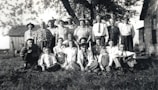 A group of people of varying ages are gathered outside, posing for a photograph. They are arranged in three rows, with some seated in the grass and others standing. Many wear vintage clothing, including patterned shirts and dresses, suggesting a setting from the mid-20th century. The backdrop includes trees and a couple of buildings, indicating a rural or suburban environment.