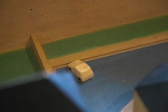 A small white toy car is placed on a multicolored surface, including green and blue hues. The environment appears to be a wooden setting, possibly part of a larger model or diorama.