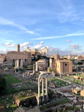A panoramic view of ancient ruins, including tall columns and scattered stone remnants in an archaeological site. In the background, historic buildings and a monument with statues enhance the scene, all under a partly cloudy blue sky.