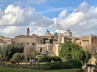 An urban landscape featuring historical architecture with a blend of classical and modern structures. There are ancient Roman ruins in the foreground surrounded by lush greenery. The skyline includes prominent historical buildings with statues and columns against a backdrop of partly cloudy skies.