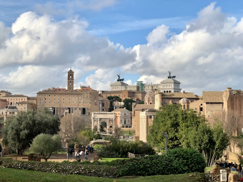 An urban landscape featuring historical architecture with a blend of classical and modern structures. There are ancient Roman ruins in the foreground surrounded by lush greenery. The skyline includes prominent historical buildings with statues and columns against a backdrop of partly cloudy skies.