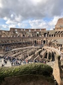 A historical amphitheater with ancient stone structures and arches. The central area is surrounded by multiple rows of spectator seating. Several people are walking and gathering around, indicating it is a tourist attraction. The sky is partly cloudy, casting a natural light over the scene.