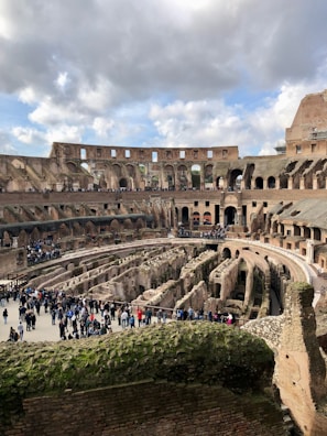 A historical amphitheater with ancient stone structures and arches. The central area is surrounded by multiple rows of spectator seating. Several people are walking and gathering around, indicating it is a tourist attraction. The sky is partly cloudy, casting a natural light over the scene.