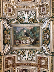 An intricately decorated ceiling in a rich baroque style featuring a painting depicting a historical or religious scene with a group of people in robes. The surrounding architecture includes ornate gold leaf detailing, relief sculptures of figures with wings, and elaborate scrollwork.