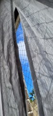 A sheer white curtain is gently blowing in front of a window with a safety mesh. Beyond the mesh, a glimpse of a bright blue sky with a few clouds is visible. A small section of a tree with green leaves is in the background.