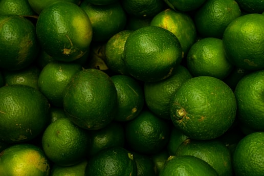 A heap of fresh green limes with a smooth texture, tightly packed together. Each lime displays a vibrant green color with patches of lighter and darker hues.