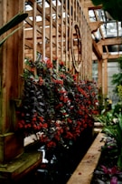 A wooden trellis structure with vibrant red flowers cascading down in an indoor garden setting. The wooden framework is detailed with a round opening, allowing light to filter through. A narrow water feature runs parallel to the structure, surrounded by lush green plants.