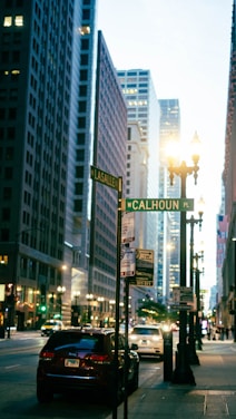 Street scene in an urban environment with tall buildings on either side. A road sign displaying 'N LaSalle St' and 'W Calhoun Pl' is visible, along with several parked cars and some passing vehicles. Bright streetlights illuminate the area, and people can be seen in the distance.