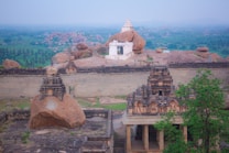 Ancient stone structures are surrounded by large rocky formations and lush greenery. A prominent white temple stands atop a massive boulder, overlooking the expansive landscape and providing a contrast to the earthy tones of the rocks and temple ruins below.