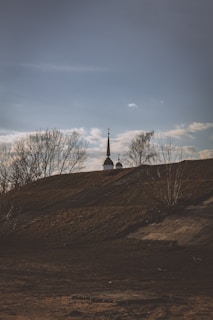 A silhouette of a church with two domes is visible against a clear blue sky. The scene includes bare trees and a grassy hill in the foreground, casting shadows as the sun sets or rises.
