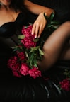 A female figure is lying on a dark leather surface, wearing black lingerie. Her hand is gently resting on a bouquet of vibrant pink peonies with lush green leaves, which are placed across her body.