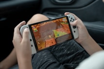 A pair of hands is holding a white handheld gaming console displaying a colorful video game. The game screen shows a vibrant scene with elements like stars and a cartoonish landscape. The person appears to be seated, as a partial view of legs in dark-colored clothing is visible in the background.