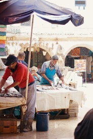 A marketplace scene with vendors working at a fish stall. Two men are handling fish spread out on tables covered with white cloth. In the background, an intricate architecture is visible with arches, and colorful textiles are displayed. An umbrella provides shade to the vendors.