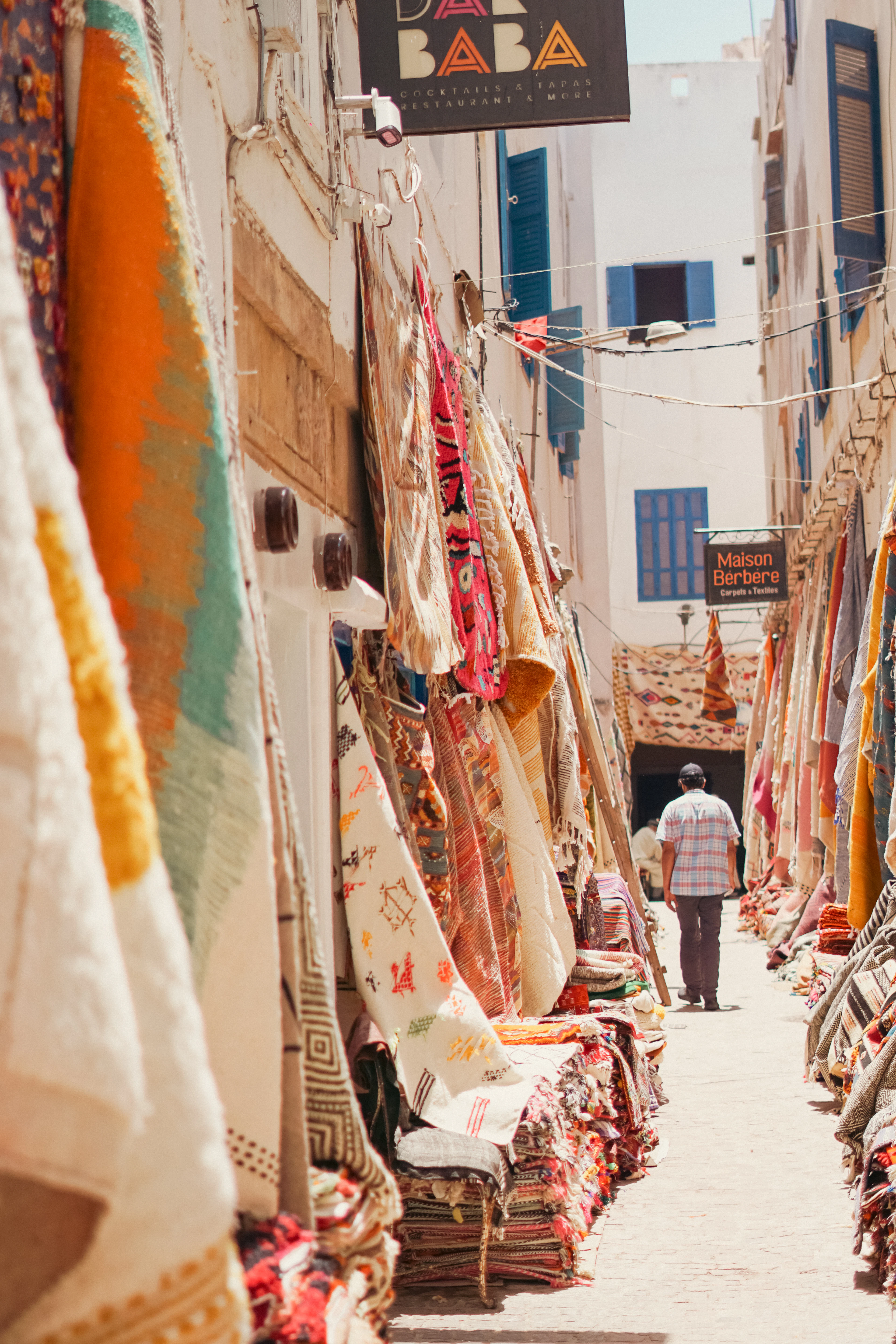 A narrow Essaouira medina alley hung with Berber rugs and carpets stacked outside the shops