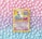 A vintage Pokémon card featuring Mew is placed against a pastel background of polka dots. The card, with a yellow border, displays an illustration of Mew with vibrant shades of pink and blue. The background features a gradient of pink, purple, and blue tones with evenly spaced white dots.