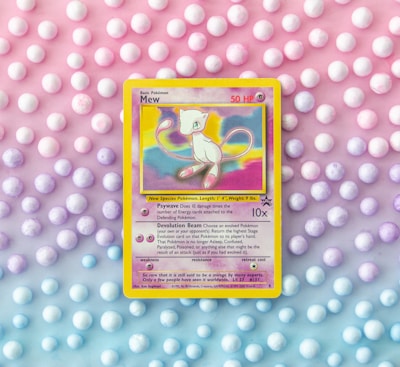 A vintage Pokémon card featuring Mew is placed against a pastel background of polka dots. The card, with a yellow border, displays an illustration of Mew with vibrant shades of pink and blue. The background features a gradient of pink, purple, and blue tones with evenly spaced white dots.