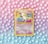 A vintage Pokémon card featuring Mew is placed against a pastel background of polka dots. The card, with a yellow border, displays an illustration of Mew with vibrant shades of pink and blue. The background features a gradient of pink, purple, and blue tones with evenly spaced white dots.