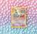A vintage Pokémon card featuring Mew is placed against a pastel background of polka dots. The card, with a yellow border, displays an illustration of Mew with vibrant shades of pink and blue. The background features a gradient of pink, purple, and blue tones with evenly spaced white dots.