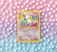 A vintage Pokémon card featuring Mew is placed against a pastel background of polka dots. The card, with a yellow border, displays an illustration of Mew with vibrant shades of pink and blue. The background features a gradient of pink, purple, and blue tones with evenly spaced white dots.