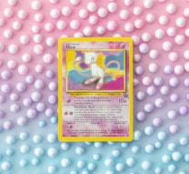 A vintage Pokémon card featuring Mew is placed against a pastel background of polka dots. The card, with a yellow border, displays an illustration of Mew with vibrant shades of pink and blue. The background features a gradient of pink, purple, and blue tones with evenly spaced white dots.