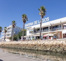 A marina lined with palm trees and a modern white building, displaying the sign 'Marina de Lagos'. The building has balconies and shops on the ground floor. People walk and sit at tables with umbrellas. A rock wall separates the walkway from the water, which is calm and reflects the clear blue sky.