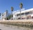 A marina lined with palm trees and a modern white building, displaying the sign 'Marina de Lagos'. The building has balconies and shops on the ground floor. People walk and sit at tables with umbrellas. A rock wall separates the walkway from the water, which is calm and reflects the clear blue sky.