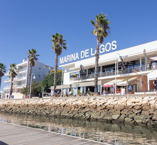 A marina lined with palm trees and a modern white building, displaying the sign 'Marina de Lagos'. The building has balconies and shops on the ground floor. People walk and sit at tables with umbrellas. A rock wall separates the walkway from the water, which is calm and reflects the clear blue sky.