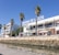 A marina lined with palm trees and a modern white building, displaying the sign 'Marina de Lagos'. The building has balconies and shops on the ground floor. People walk and sit at tables with umbrellas. A rock wall separates the walkway from the water, which is calm and reflects the clear blue sky.