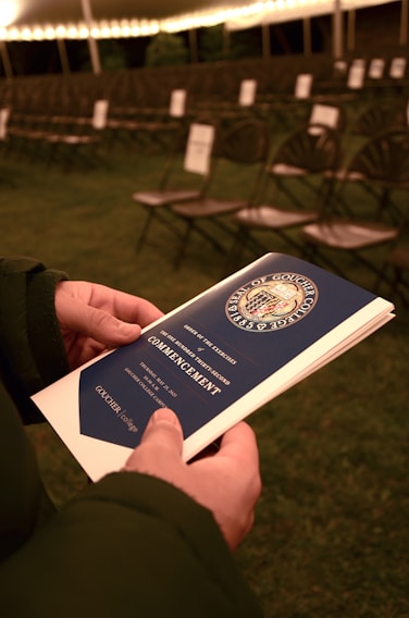 A person is holding a commencement program booklet with a blue cover featuring the seal, under a canopy with rows of empty chairs lined up in the background. The lighting suggests an evening or night setting, creating a warm atmosphere.
