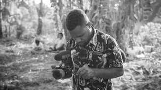A man is focusing on a camera, which is equipped with a microphone, amidst a natural, wooded setting. Other individuals are sitting in the background, surrounded by tall trees and foliage, suggesting a secluded or forested environment. The lighting is soft, giving a tranquil atmosphere to the scene.