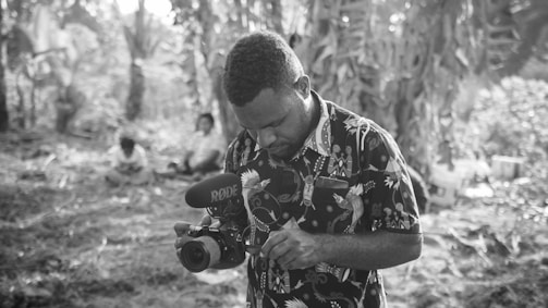 A man is focusing on a camera, which is equipped with a microphone, amidst a natural, wooded setting. Other individuals are sitting in the background, surrounded by tall trees and foliage, suggesting a secluded or forested environment. The lighting is soft, giving a tranquil atmosphere to the scene.