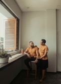A man and a woman are inside a room with light gray walls, sitting near a window with wooden blinds. They are both dressed in matching rust-colored tops, with the man seated on a stool and the woman standing beside him, resting her hand on his shoulder. The room has potted plants along the windowsill, and the mood appears warm and intimate.