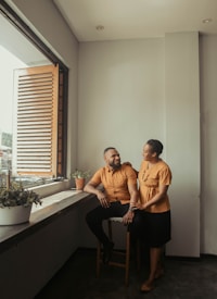 A man and a woman are inside a room with light gray walls, sitting near a window with wooden blinds. They are both dressed in matching rust-colored tops, with the man seated on a stool and the woman standing beside him, resting her hand on his shoulder. The room has potted plants along the windowsill, and the mood appears warm and intimate.