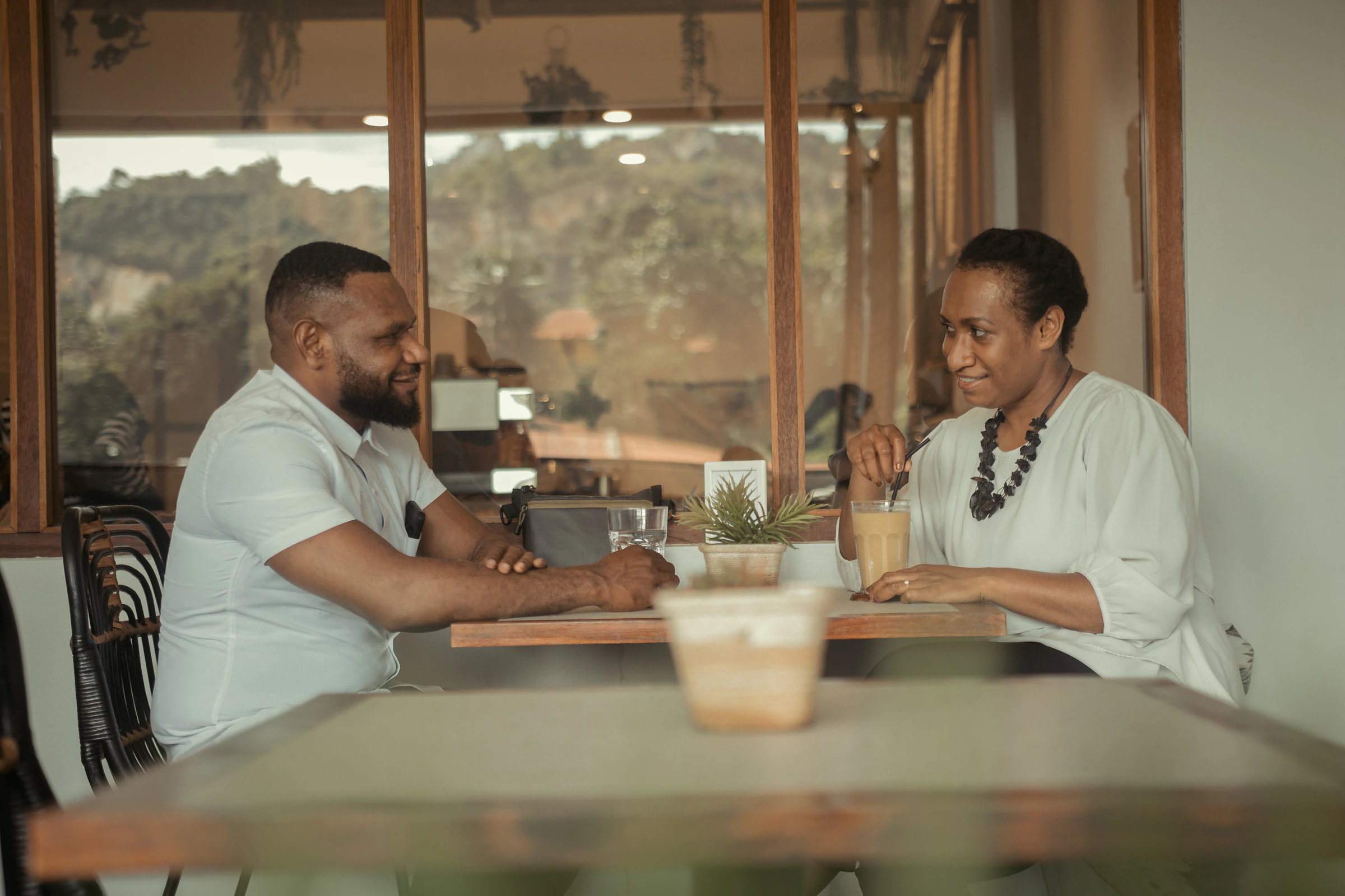 A woman in a pale green blouse smiling mid-conversation across a timber table from a man in a white shirt, beside a window looking onto trees