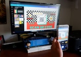 A person is holding a smartphone in front of a computer monitor, capturing a picture of a screen displaying a design with a QR code. The design is colorful with checkerboard patterns and includes text encouraging users to scan the QR code. The desk is cluttered with books, a small figurine, and office supplies.