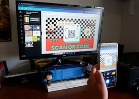 A person is holding a smartphone in front of a computer monitor, capturing a picture of a screen displaying a design with a QR code. The design is colorful with checkerboard patterns and includes text encouraging users to scan the QR code. The desk is cluttered with books, a small figurine, and office supplies.