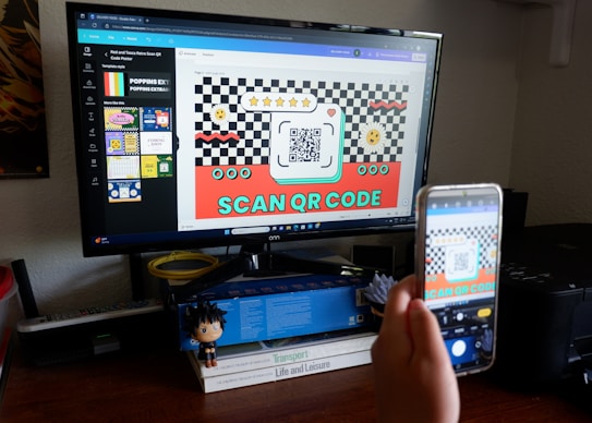 A person is holding a smartphone in front of a computer monitor, capturing a picture of a screen displaying a design with a QR code. The design is colorful with checkerboard patterns and includes text encouraging users to scan the QR code. The desk is cluttered with books, a small figurine, and office supplies.
