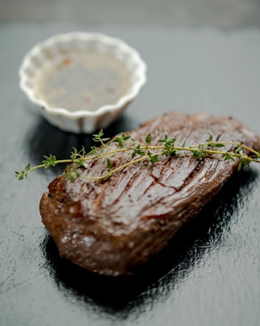 A cooked steak rests on a dark, glossy surface, garnished with a sprig of fresh thyme. In the background, a small white, fluted bowl holds a dipping sauce.
