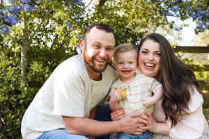 A cheerful family, consisting of two adults and a child, posing together outdoors surrounded by greenery and vibrant purple flowers.