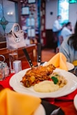 A plate of roasted pork knuckle is served with a side of mashed potatoes, sauerkraut, and parsley. Two knives are inserted into the meat. The setting is a cozy restaurant with warm lighting, wooden interior, and a decorative stained glass window. In the background, people are engaged in conversation at their table.