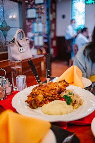 A plate of roasted pork knuckle is served with a side of mashed potatoes, sauerkraut, and parsley. Two knives are inserted into the meat. The setting is a cozy restaurant with warm lighting, wooden interior, and a decorative stained glass window. In the background, people are engaged in conversation at their table.