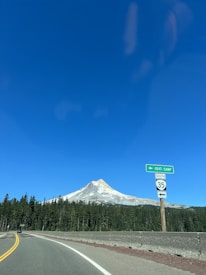 A scenic landscape of a snow-capped mountain under a clear blue sky. A two-lane road curves into the distance, bordered by dense evergreen trees. A road sign indicates directions to Government Camp and highway 35 south.