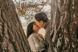 The couple sharing a gentle kiss surrounded by tall green trees and soft sunlight.