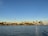 A city skyline with modern skyscrapers and the prominent Space Needle against a clear blue sky. The view is from across a body of water with gentle ripples reflecting the sunlight.