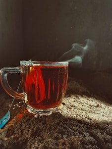 A glass cup filled with dark tea sits on a textured, earthy surface. Steam rises gently from the cup, indicating warmth. Sunlight casts a soft glow across the surface, highlighting the rich color of the tea. A tea bag tag is visible next to the cup.