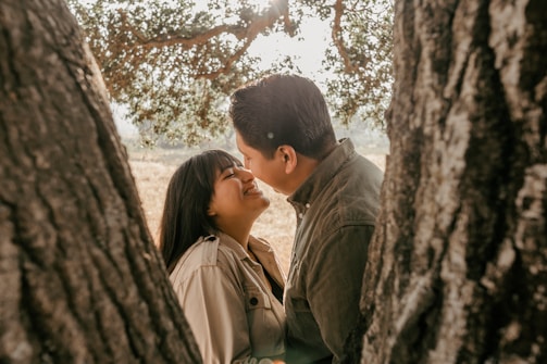 A couple is sharing a joyful and intimate moment between two tree trunks. The woman is smiling brightly as she looks at her partner. Sunlight filters through the leaves of the tree, creating a warm and tranquil atmosphere.
