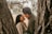 A couple is sharing a joyful and intimate moment between two tree trunks. The woman is smiling brightly as she looks at her partner. Sunlight filters through the leaves of the tree, creating a warm and tranquil atmosphere.